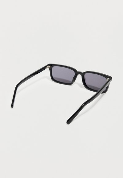 Μαύρα ορθογώνια γυαλιά ηλίου με σκουρόχρωμους φακούς που ακουμπούν σε μία λευκή επιφάνεια, εμφανιζόμενα από την πίσω πλευρά.