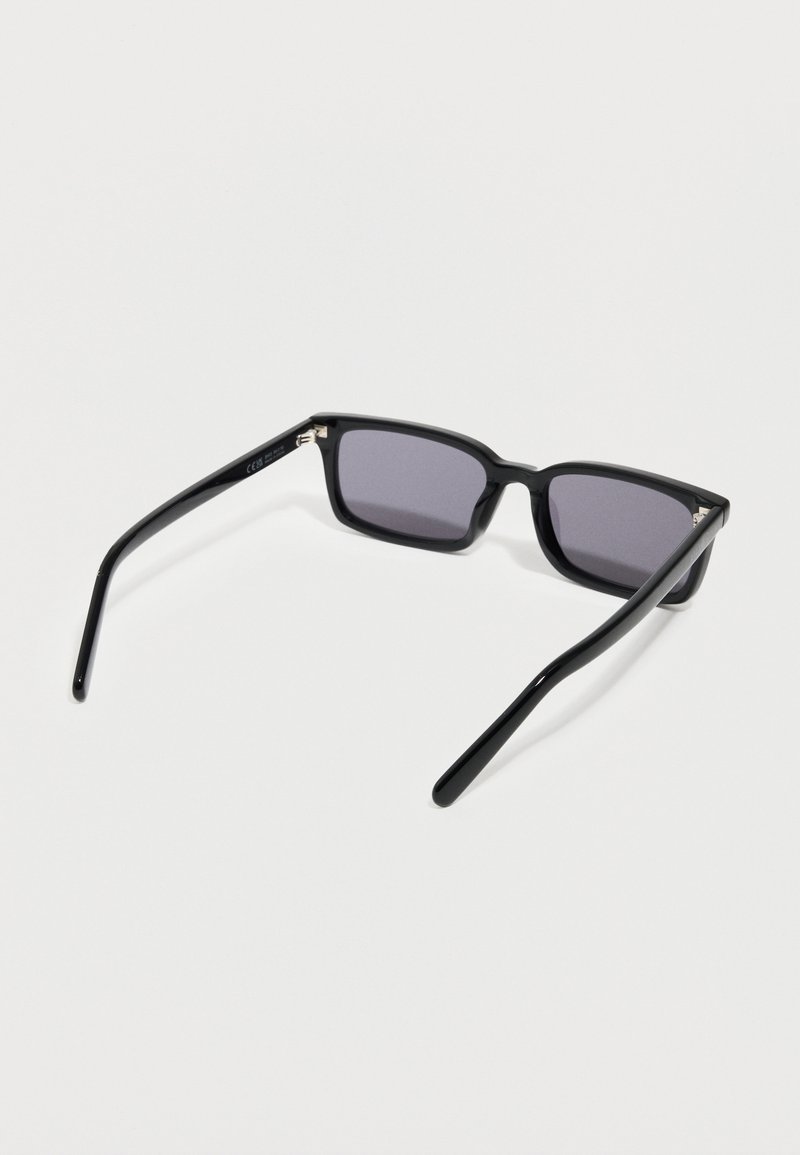 Occhiali da sole rettangolari neri con lenti scure appoggiati su una superficie bianca, mostrati dal retro.