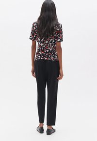 Schwarzes Blusenshirt mit kurzen Ärmeln und roten sowie weißen Mustern, kombiniert mit schwarzen, taillierten Hosen und schwarzen flachen Schuhen. Rückansicht.