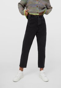 Jeans de mezclilla negra de cintura alta con corte recto, que cuentan con cinco bolsillos y un dobladillo deshilachado, combinados con zapatillas blancas de caña alta.