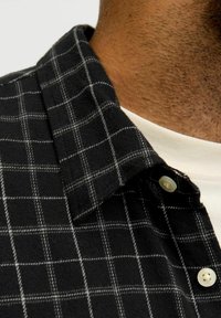 Lähivõte inimese kaelast ja ülemisest rinnast, kes kannab musta ruudulist särke valge ümardatud kaelusega T-särgi peal.