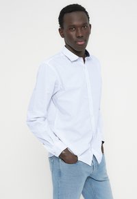 Ανοιχτό γαλάζιο πουκάμισο με μικρό σχεδιαστικό μοτίβο, εφαρμοστή γραμμή, σκουρόχρωμο γιακά σε αντίθεση και μακρυά μανίκια. Φορεμένο με ανοιχτόχρωμο τζιν παντελόνι.