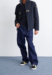 Marine vindjakke med lynlås-lommer, båret over en sort skjorte. Blå brede jeans og sorte sneakers med gule detaljer fuldender looket.