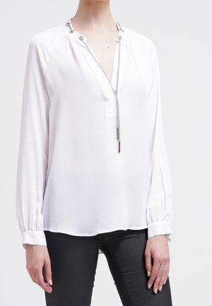 Hvid bluse med lange ærmer og V-hals med sølvkæde ved halsudskæringen, båret med sorte bukser.