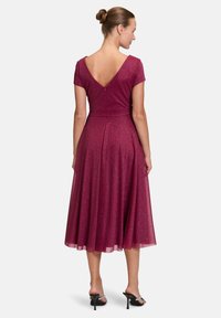 Bordeauxrotes Kleid mit kurzen Ärmeln, V-Ausschnitt und schwingendem Rock. Der Stoff hat Glitzer, mit einem versteckten Reißverschluss im Rücken. Knielange Länge.