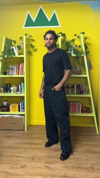 Svart t-shirt med korta ärmar och randiga vida byxor. Står på trägolv framför gröna hyllor fyllda med växter och böcker mot en gul vägg.
