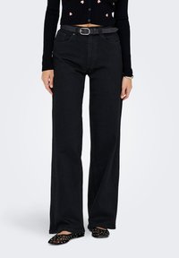 Femme portant un jean noir taille haute et large avec une ceinture noire, un pull court noir, des bagues, un bracelet et des ballerines noires à clous.