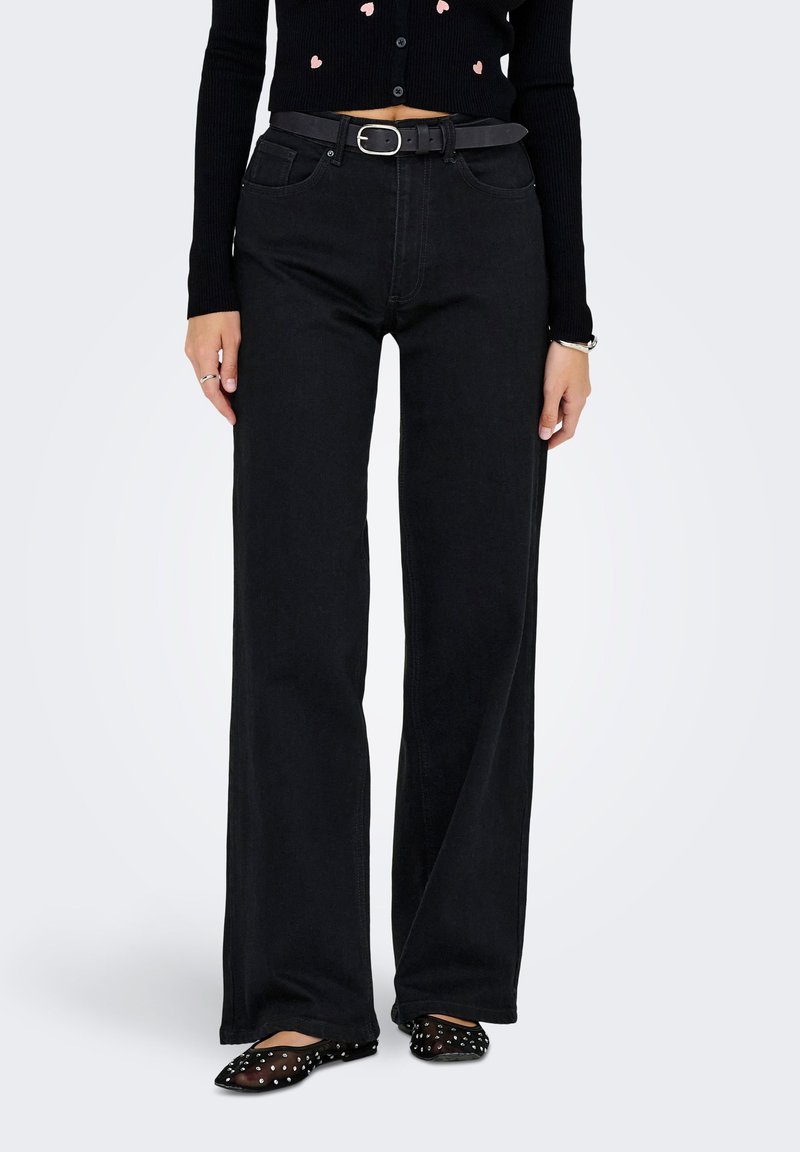 Femme portant un jean noir taille haute et large avec une ceinture noire, un pull court noir, des bagues, un bracelet et des ballerines noires à clous.