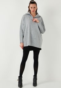 Pulover oversized gri deschis cu guler înalt, textură cu rigole și o croială lejeră, asociat cu colanți negri și cizme până la glezne.