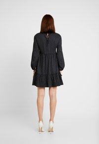 Vestido negro con lunares, de mangas largas, cintura ceñida y dobladillo con volantes en capas. Detalle de abertura en la espalda y mangas ajustables con lazo.