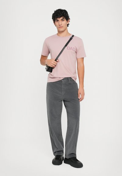 Lys rosa t-skjorte med korte ermer og teksten "DAD", kombinert med grå kordfløyelsbukser og svarte joggesko. Modellen holder en liten svart veske.