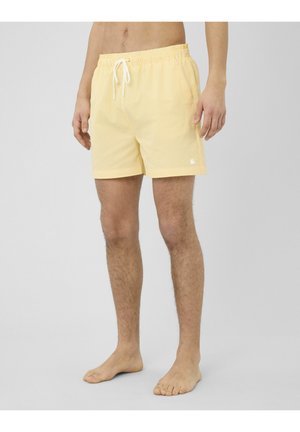 Homme portant un short de bain à cordon de serrage rayé jaune et blanc, debout pieds nus sur un fond neutre.