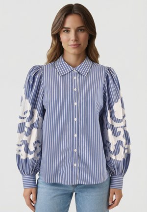 Femme portant un chemisier à rayures verticales bleu et blanc avec des manches bouffantes ornées de broderies florales blanches, associé à un jean bleu clair.