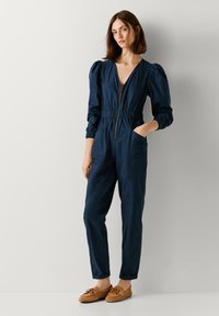 Ολόσωμη φόρμα σε ναυτικό μπλε χρώμα με φερμουάρ μπροστά, ελαστική μέση, puff μανίκια και πλαϊνές τσέπες. Συνδυάζεται με ταμπά παπούτσια χωρίς κορδόνια.