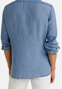 Personne portant une chemise bleu clair à manches longues retroussées et un pantalon blanc, vue de dos.