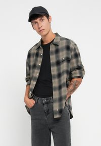 Camisa de franela a cuadros verdes y negros con mangas largas, botón en la parte delantera, bolsillo en el pecho, combinada con una camiseta negra y jeans gris oscuro.