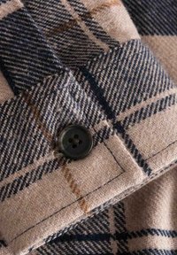 Tessuto di lana a quadri con tonalità blu navy, beige e marrone. Un bottone nero a quattro fori è cucito sul tessuto, evidenziando dettagli di cucitura raffinati.