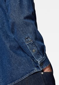 Denim shirt met lange mouwen, donkerblauwe kleur, gedetailleerde stiksels, knoopmanchet met metalen hardware en een zichtbare zijzak.
