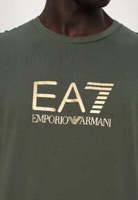 Olivengrønn bomulls-T-skjorte med et stort gulllogo med teksten "EA7 EMPORIO ARMANI." Kort ermer og rund hals.