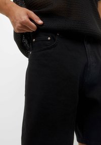 Pantalones cortos de mezclilla negra con una textura suave, que presentan remaches metálicos y un bolsillo frontal. La tela es opaca, con un diseño de corte recto.