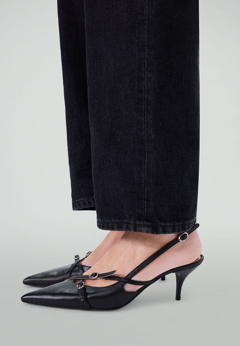 Talons aigus en cuir noir avec un design à dos ouvert, présentant un design découpé et des accents en métal argenté, reposant sur un petit talon aiguille.