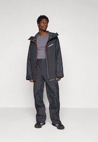 Must veekindel jope tekstureeritud varrukatega, oranžide aktsentidega, ja püksid geomeetrilise mustriga. Kanditud halli aluskihti ja mustade jalanõudega.