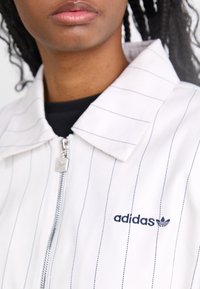 Bílá bunda na zip s tmavě modrými proužky a límcem, který lze ohrnout dolů. Na levé straně je vyšitá logo Adidas.