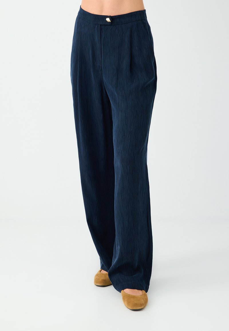 Pantalon bleu marine texturé à jambes droites avec plis avant et un petit bouton rond doré à la taille, porté avec des chaussures plates marron clair.