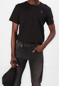 Camiseta de algodón negra con cuello redondo y un pequeño logo, combinada con jeans de mezclilla gris oscuro con bolsillos frontales y herrajes negros.