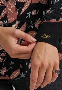 Camiseta preta com padrão floral em castanho e branco, apresentando um "LW" bordado a ouro na manga. Mão a ajustar a manga.