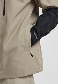 Giacca impermeabile beige con maniche nere, dotata di tasca con zip e tessuto testurizzato. Dettaglio del logo sulla manica.