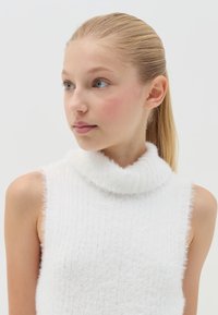 Suéter sin mangas de cuello alto en color blanco, confeccionado en un tejido suave y texturizado, con un diseño acanalado y un cuello esponjoso.
