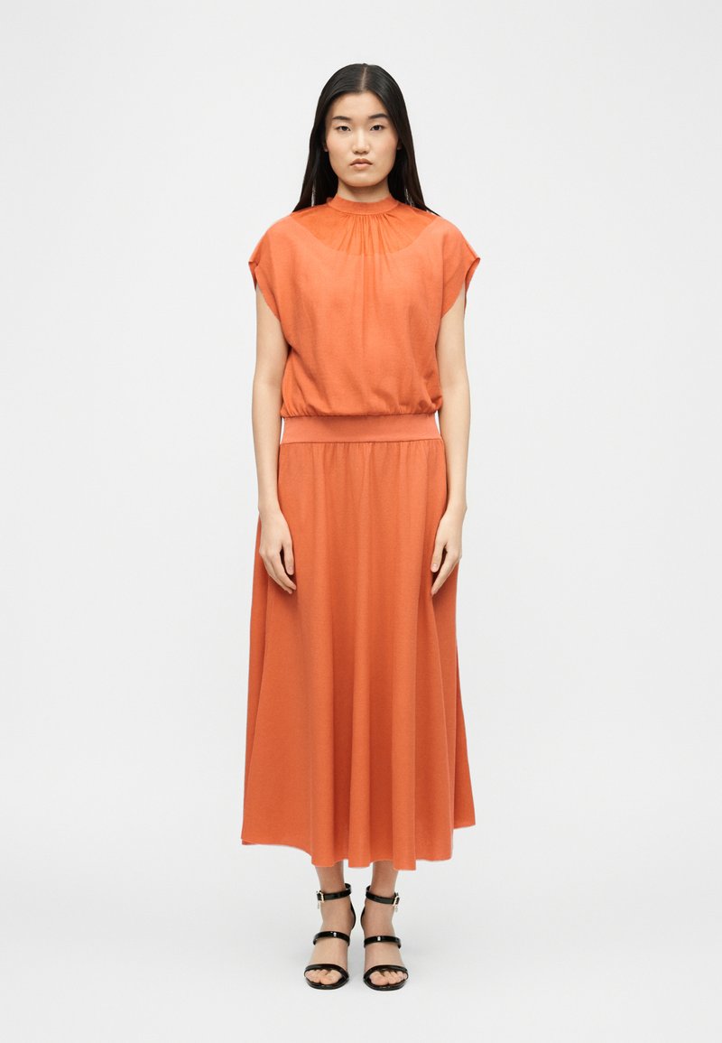 Femme debout portant un chemisier orange à manches courtes avec un col montant et une jupe longue assortie, accompagnée de sandales noires à lanières.