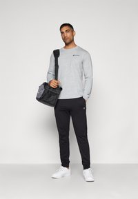 Grå langermet skjorte med logo, svarte joggebukser og hvite sneakers. Bærer en svart og grå bag med stropper.