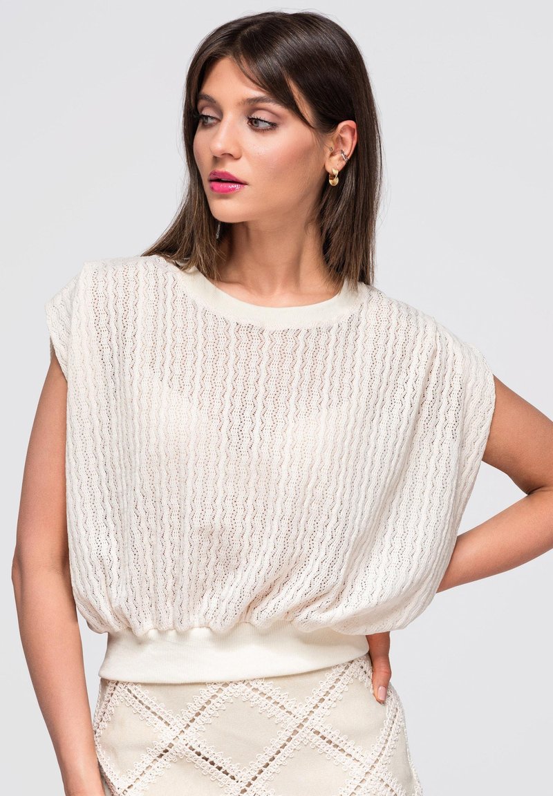 Femme portant un haut en maille blanc ample et une jupe texturée assortie, regardant vers sa gauche avec une main sur la hanche sur un fond uni.