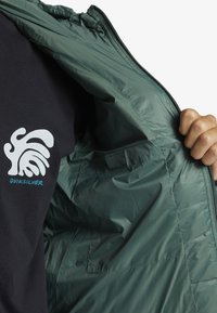 Green puffer jacket with a smooth, shiny texture. Interior features a contrasting black t-shirt with a white wave logo and blue brand text.