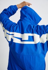 Kék kapucnis dzseki, amelynek hátulján nagy fehér grafikai szöveg található. Az anyag könnyűnek tűnik, sima tapintású és elasztikus mandzsettákkal rendelkezik.