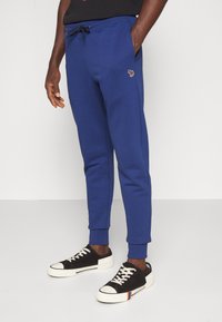 Man wearing blue jogger pants with drawstring waist and black low-top sneakers with white soles and laces, standing with one hand in pocket.