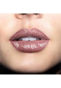 Rouge à lèvres mauve mat sur des lèvres pulpeuses avec une finition brillante, mettant en valeur une texture lisse et un contour défini. Dents visibles entre les lèvres.