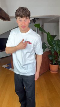 Νεαρός άνδρας με λευκό μπλουζάκι Adidas που δείχνει το λογότυπο, στέκεται σε ένα σαλόνι με ξύλινο πάτωμα, μεγάλο γλαστράκι και καναπέ στο φόντο.