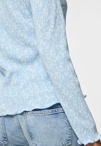 Maglietta a maniche lunghe a righe blu chiaro con motivo floreale bianco. Presenta un orlo e polsini ondulati. Abbinata a jeans chiari in denim.