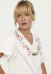 T-shirt blanc avec un col en V et des manches courtes à volants, ornée de broderies florales roses autour du col et de motifs en losange décoratifs sur le corps.