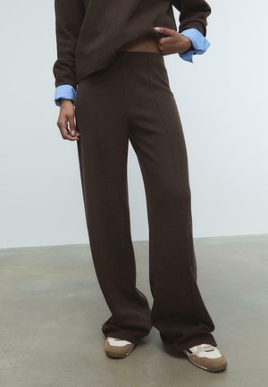 Personne portant un pantalon marron à jambes larges, un haut marron avec des poignets de chemise bleus et des baskets blanches, debout sur un sol gris.