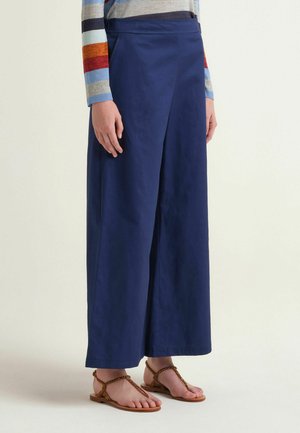 Personne portant un pantalon ample bleu marine, une chemise rayée à manches longues et des sandales plates marron, debout sur un fond uni.