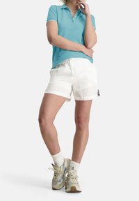 Polo shirt turquoise avec un col, associé à un short blanc. Les accessoires incluent des baskets beiges avec des accents bleus et des chaussettes blanches.