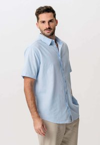 Mand med kort, krøllet hår og skæg, iført en lyseblå, kortærmet skjorte med knapper og beige bukser, der står foran en ensfarvet baggrund.