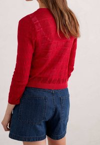 Roter Strickpullover mit strukturierter Musterung, langen Ärmeln und einem gerippten Saum, kombiniert mit blauen Denim-Shorts mit zwei Gesäßtaschen.