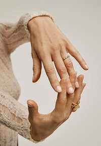 Anillo de oro con acento de turquesa en una mano; la otra mano muestra uñas naturales. Manga de suéter texturizado de color claro en el fondo.