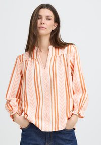 Femme portant un chemisier à rayures verticales orange et blanches à manches longues, avec un col en V et des poignets élastiques, les mains dans les poches en denim bleu.