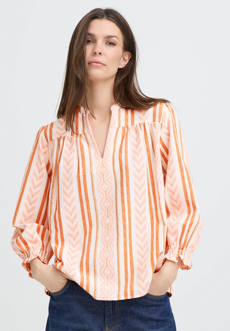 Femme portant un chemisier à rayures verticales orange et blanches à manches longues, avec un col en V et des poignets élastiques, les mains dans les poches en denim bleu.